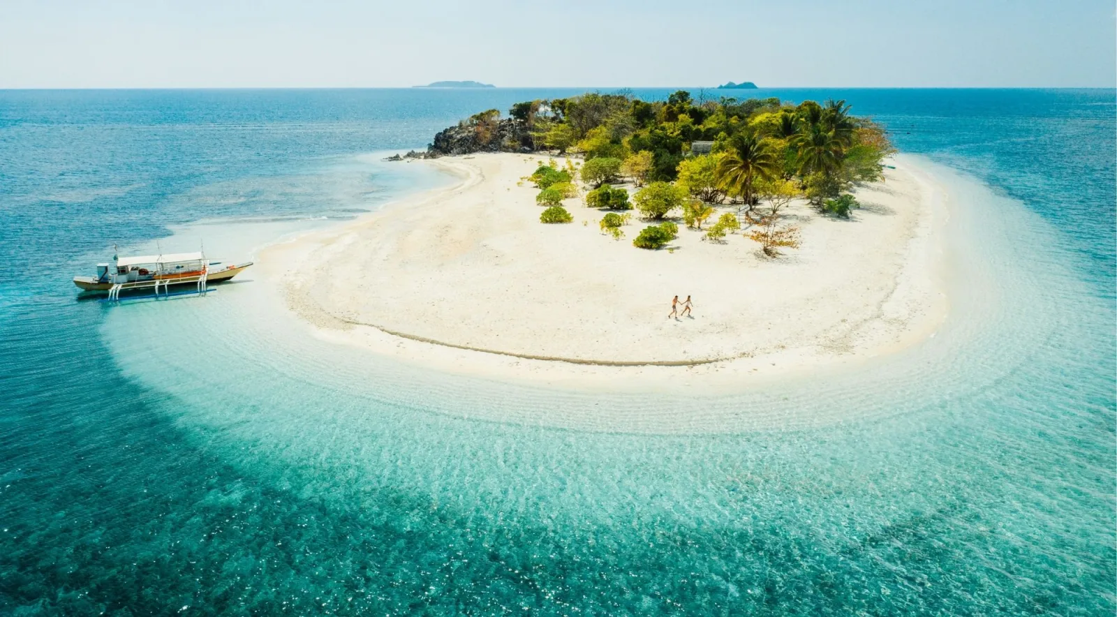 Aerial view of a pristine white sand island (sandbar) surrounded by clear turquoise water, with a couple walking and a banca anchored nearby