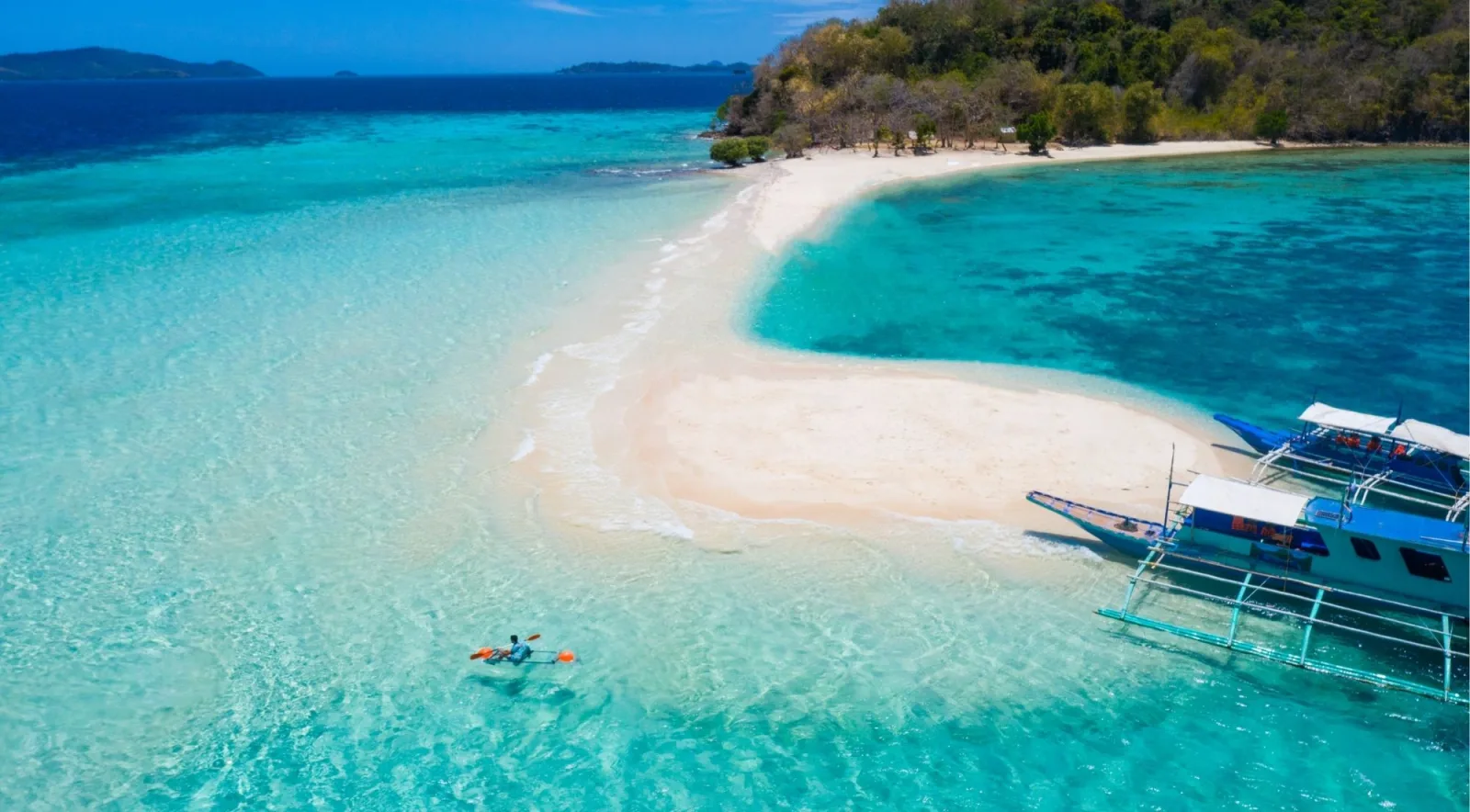 Aerial view of a curved white sandbar with a kayaker in clear water and a blue-roofed banca anchored at the tip, Palawan