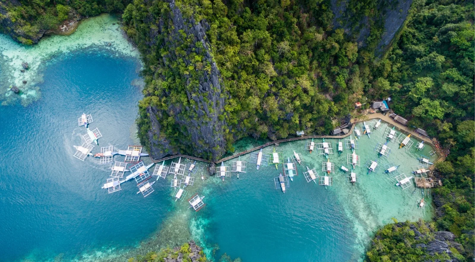 Aerial view of a fleet of traditional white outrigger bancas moored in a cove beside towering limestone karst formations, Coron