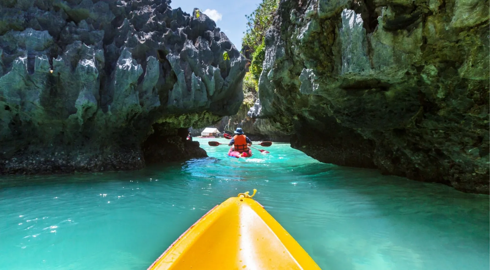 Two kayakers paddling through a narrow rocky sea tunnel with brilliant turquoise water in El Nido, Palawan
