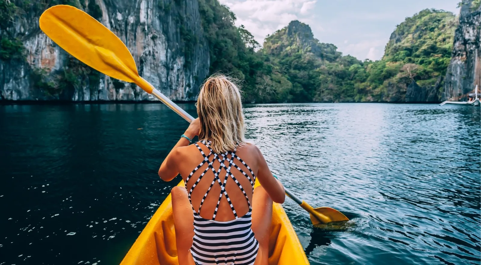 Woman paddling a yellow kayak on calm dark blue water with towering limestone karst cliffs surrounding her, El Nido